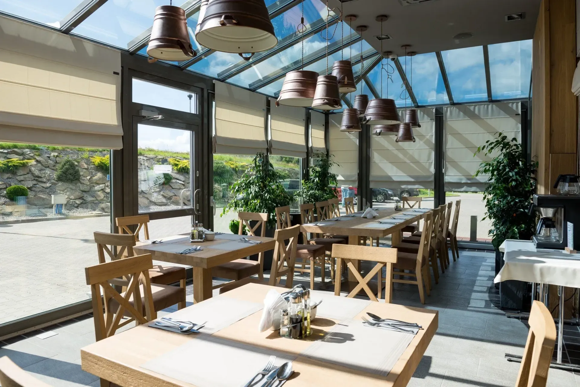 Restaurant-Innenraum mit Holztischen und -stühlen unter einem Glasdach mit Blick auf einen Garten.