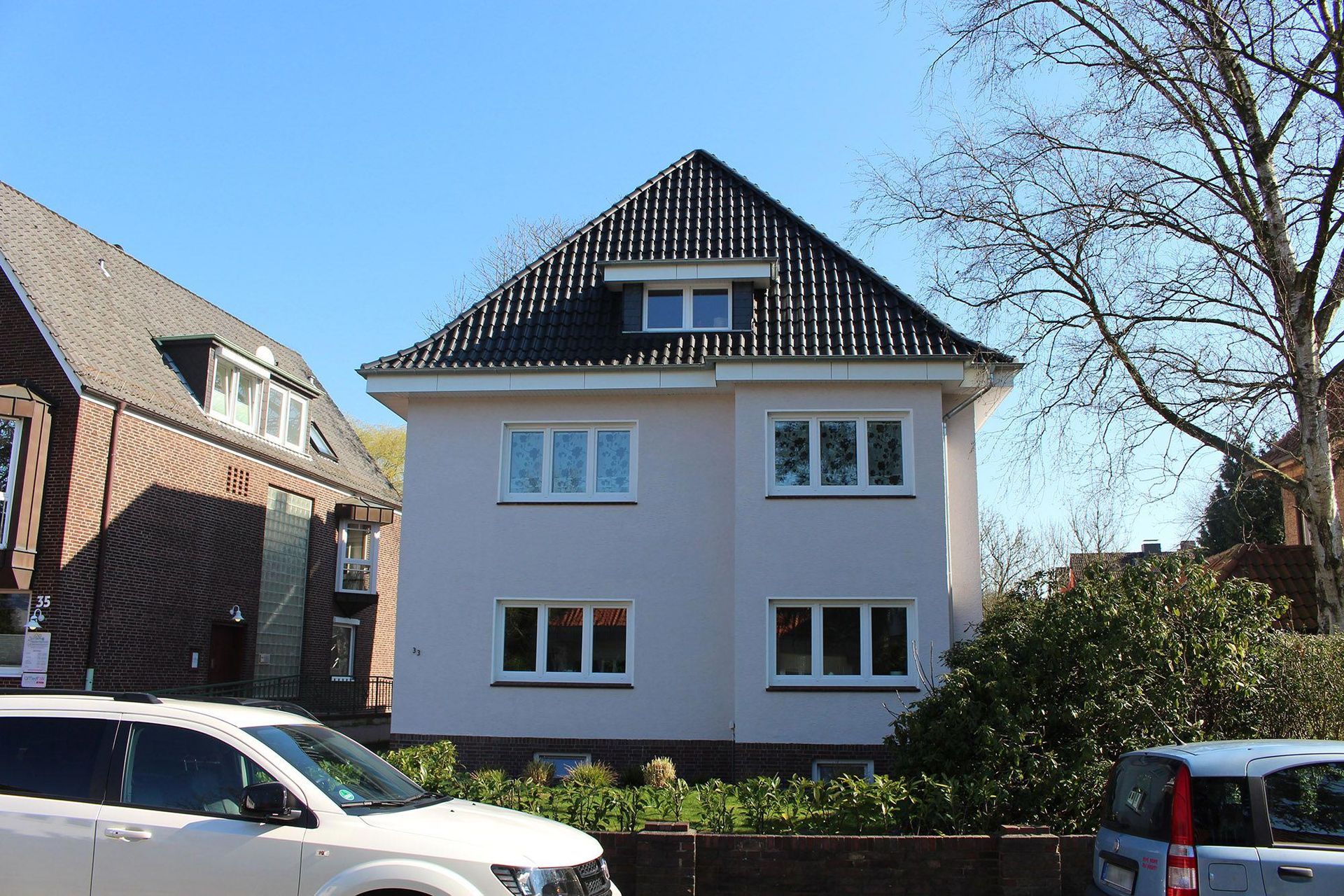 Zweistöckiges Haus mit hellen Wänden, dunklem Dach und mehreren Fenstern; im Vordergrund geparkte Autos und Bäume.