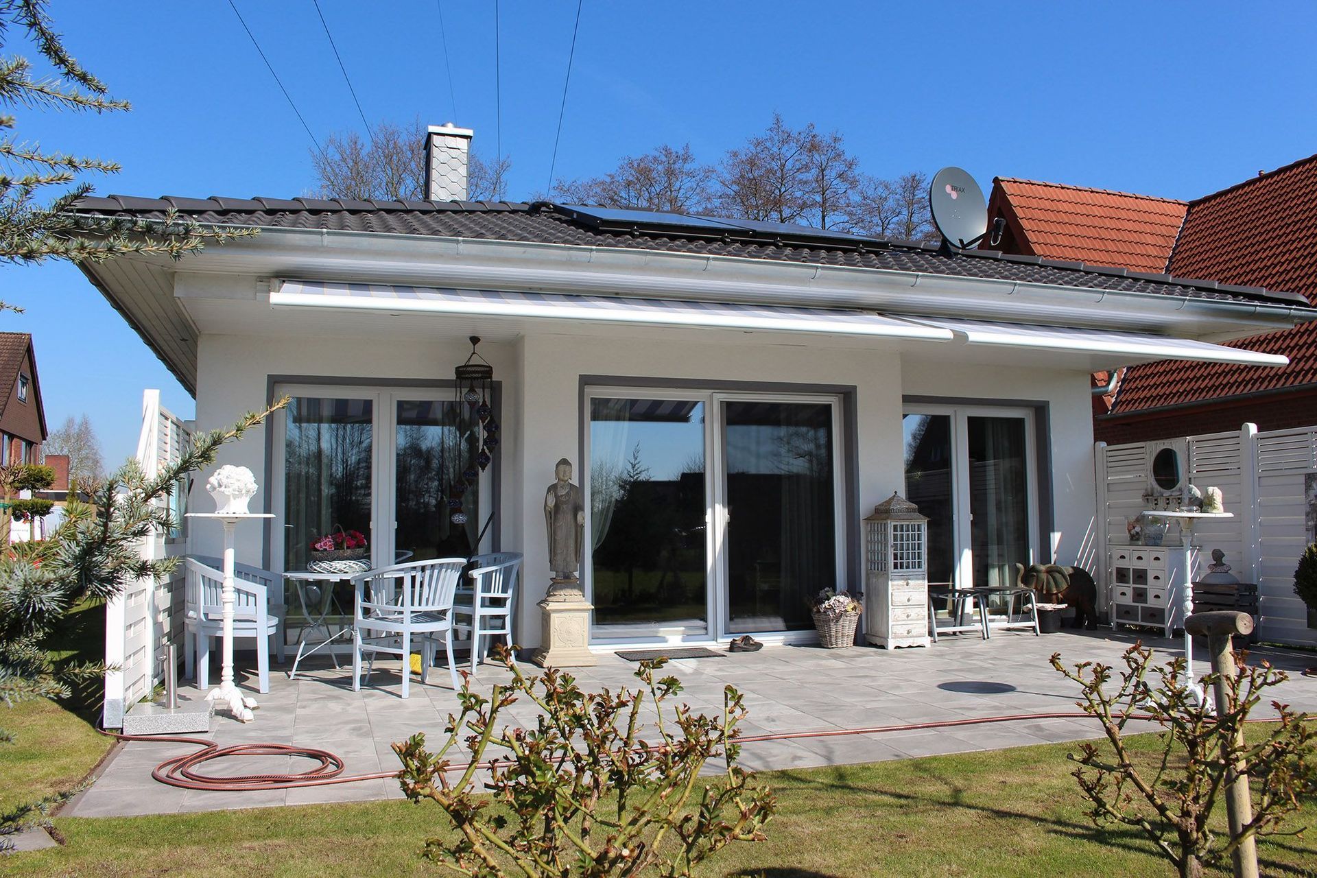 Weißes Haus mit Terrasse, Stühlen und Garten unter blauem Himmel.