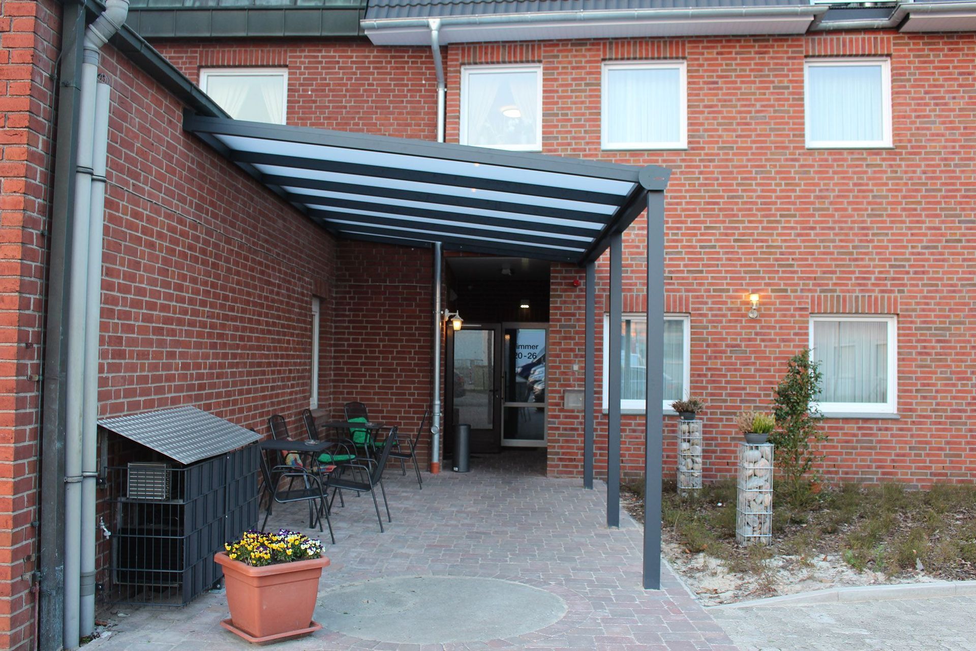 Backsteingebäude mit Vordach, Außensitzplatz und Tür, Pflanzen in Töpfen.