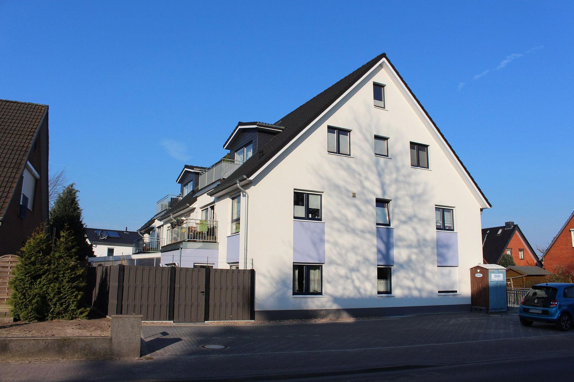 Weißes Wohnhaus mit dunklem Dach, blauen Akzenten und einem Holzzaun unter einem klaren blauen Himmel.