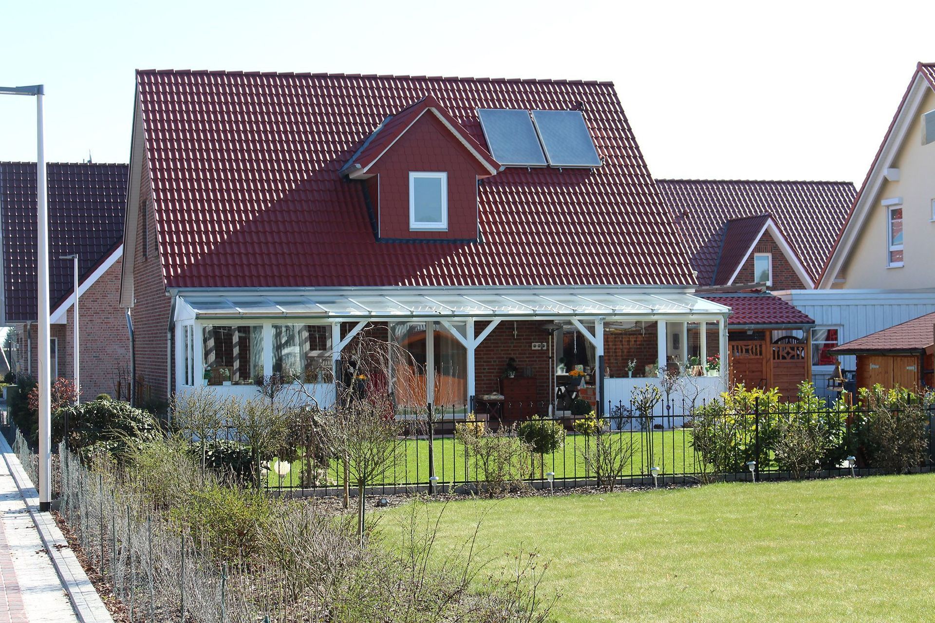 Haus mit rotem Ziegeldach, Solarpanel und Glasveranda. Sonniger Tag, grüner Rasen.