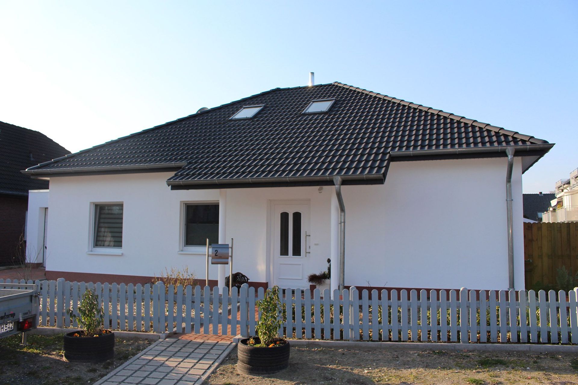 Weißes Bungalowhaus mit schwarzem Dach, kleinem Lattenzaun und zwei Topfpflanzen.