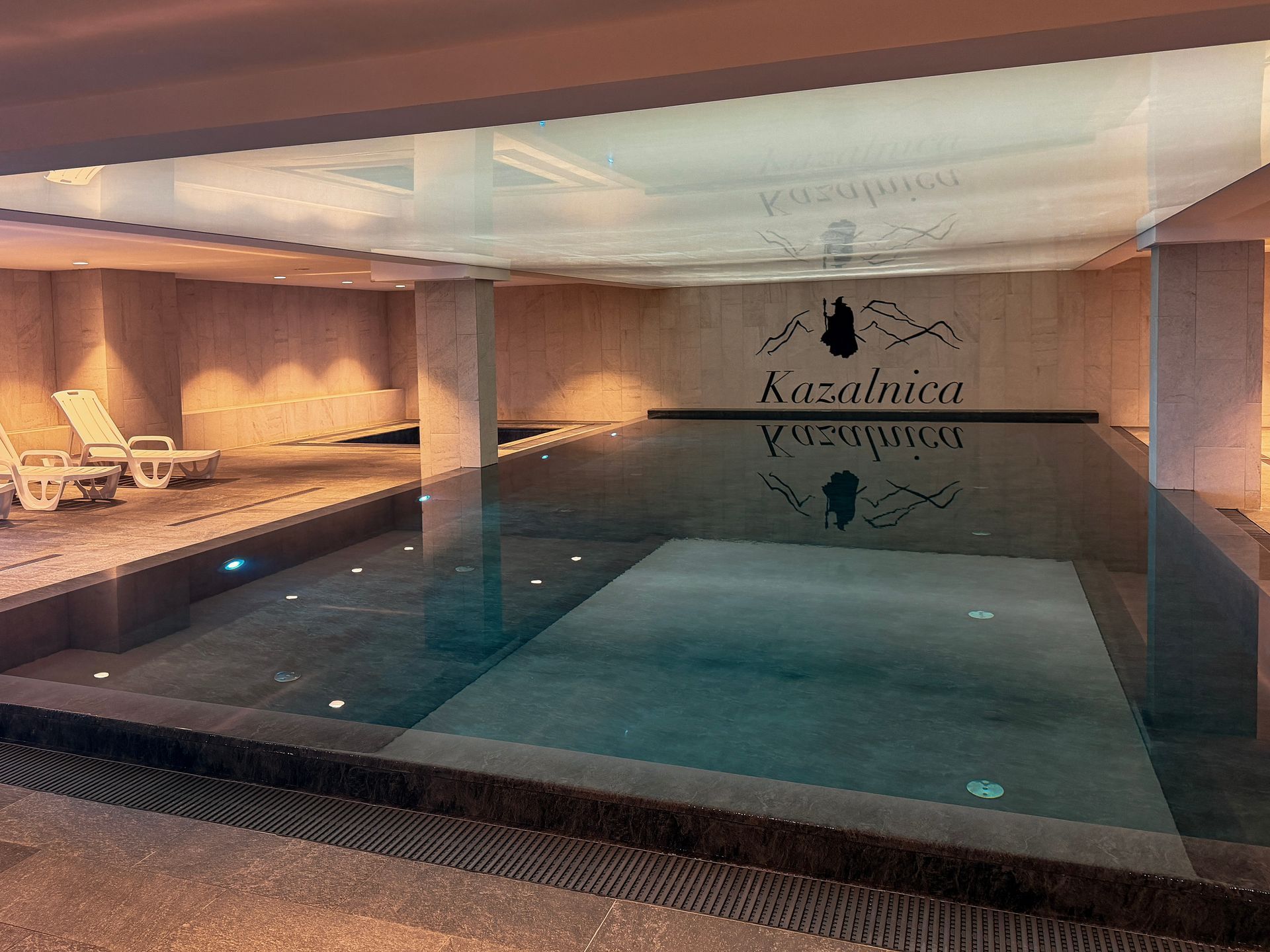 Kryty basen z spokojną, błękitną wodą, leżakami i delikatnym oświetleniem w nowoczesnym otoczeniu spa