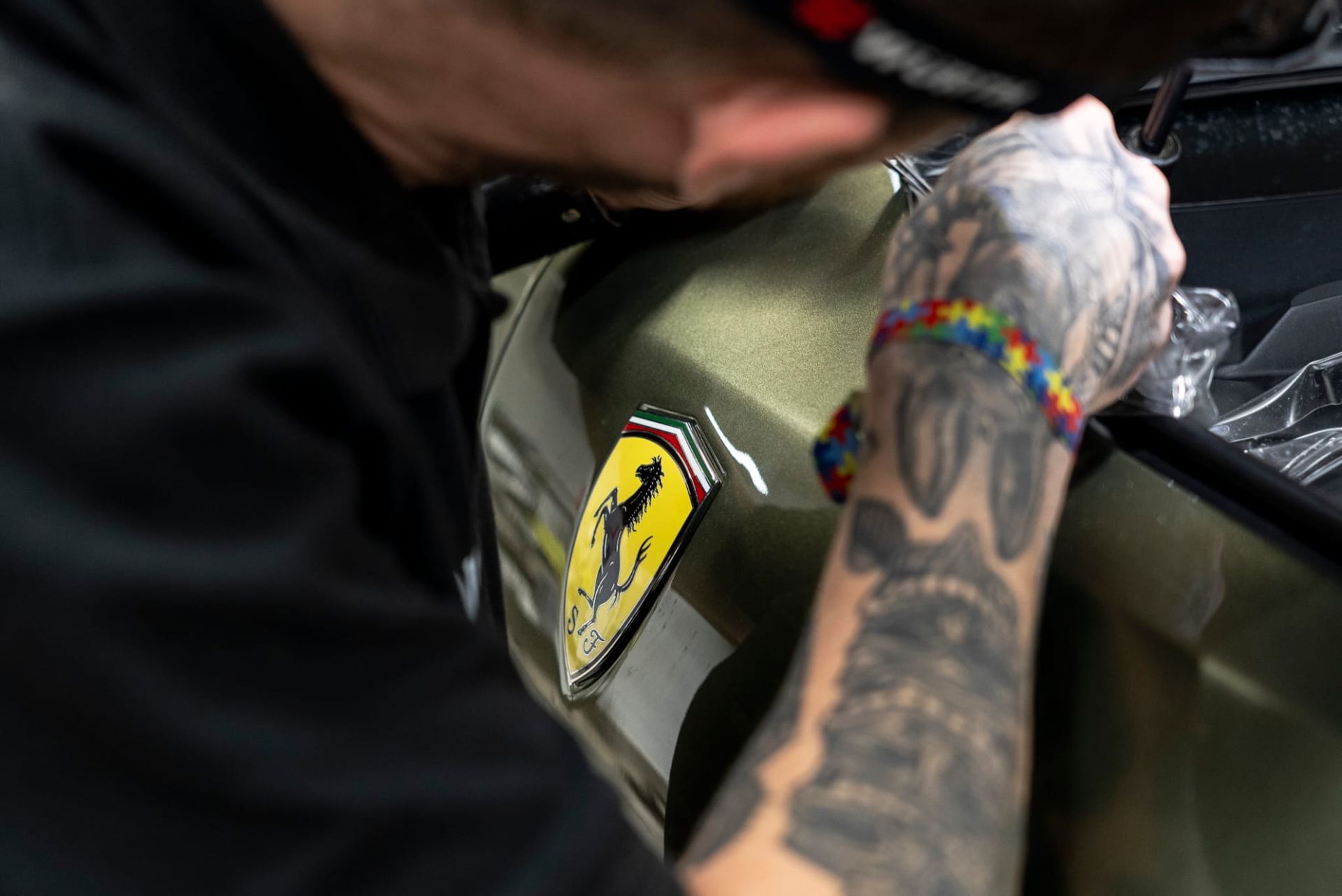 Persona aplicando detalles al emblema de Ferrari en un coche verde. Se ven tatuajes en el brazo y una pulsera de arcoíris.