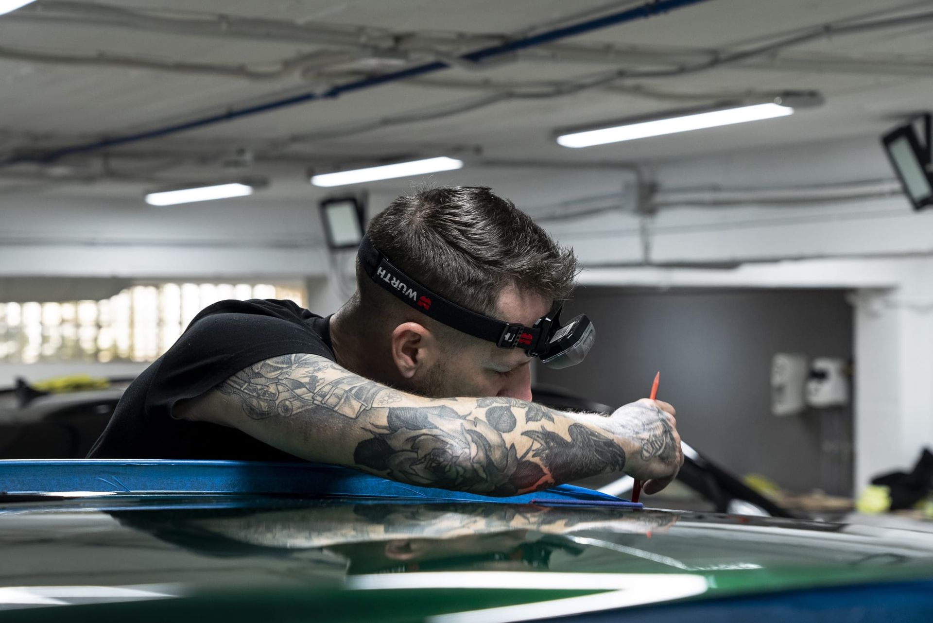 Un hombre con un faro se inclina sobre un coche, usando una herramienta. Tiene tatuajes. En un taller.