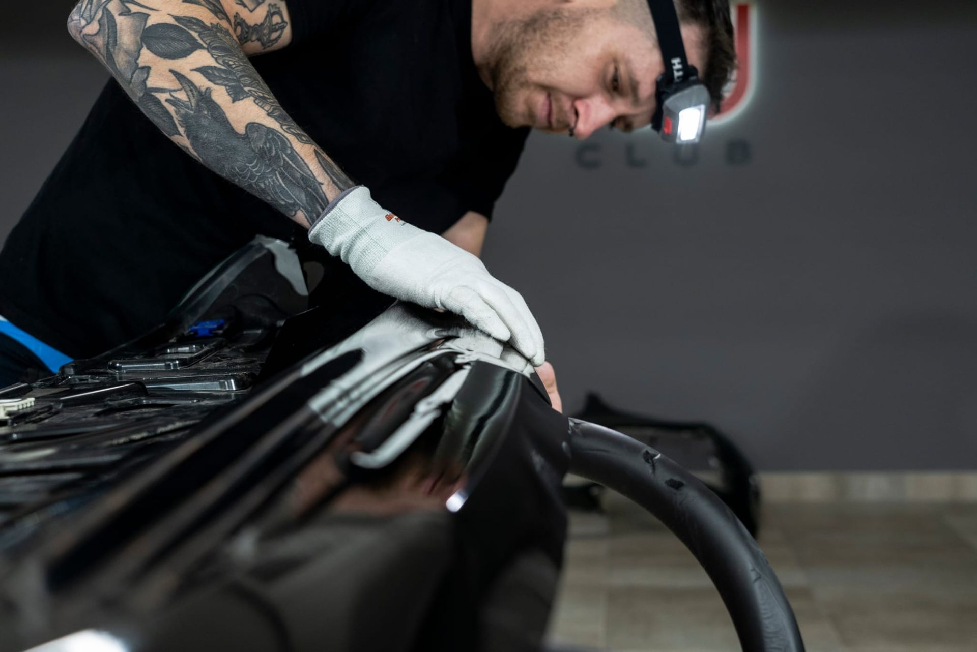 Hombre con tatuajes, con faro, trabajando en el parachoques de un coche negro en un garaje, usando una mano enguantada.