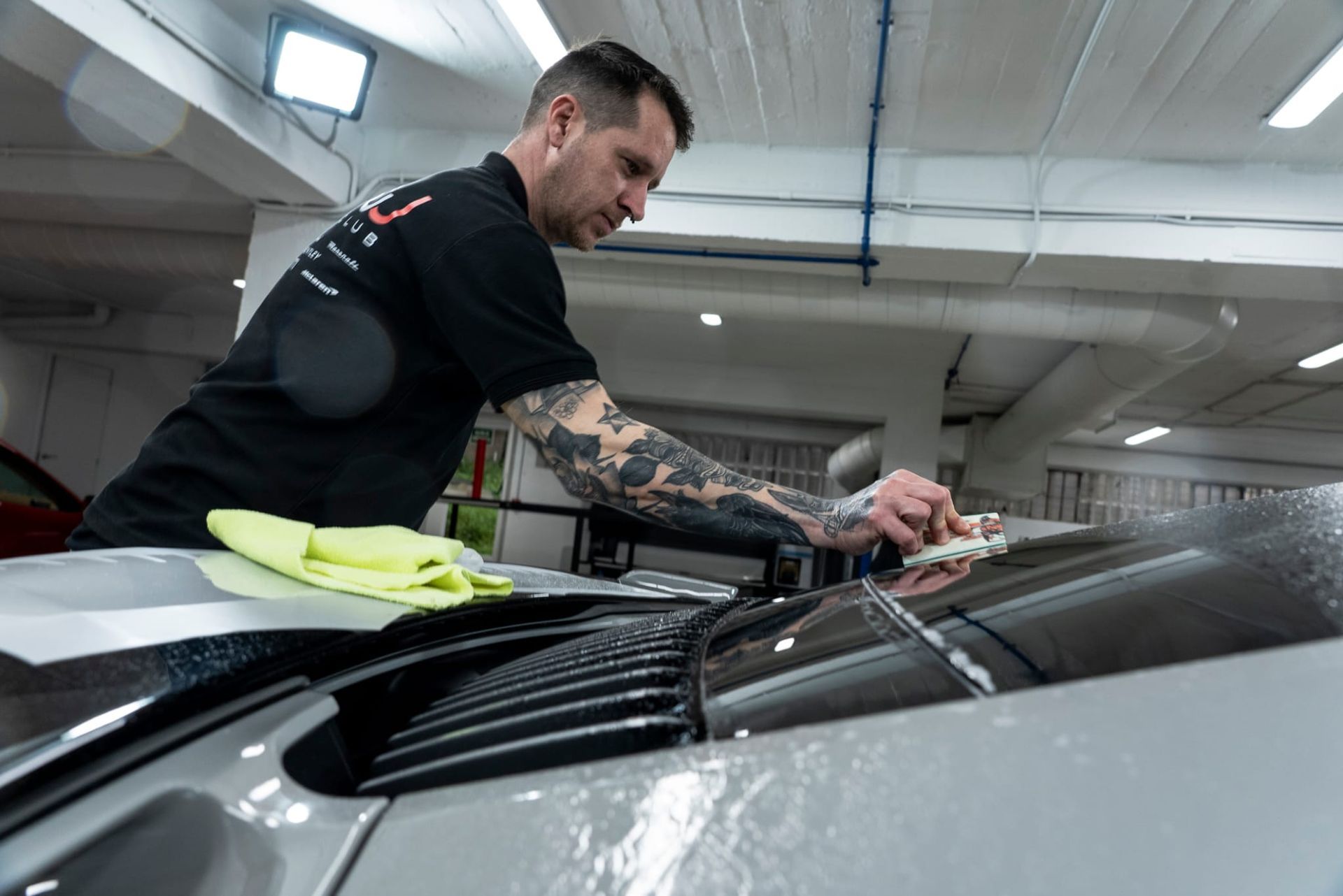 Un hombre aplica película a la ventanilla de un coche en un garaje. Lleva una camisa negra y tiene tatuajes en el brazo.