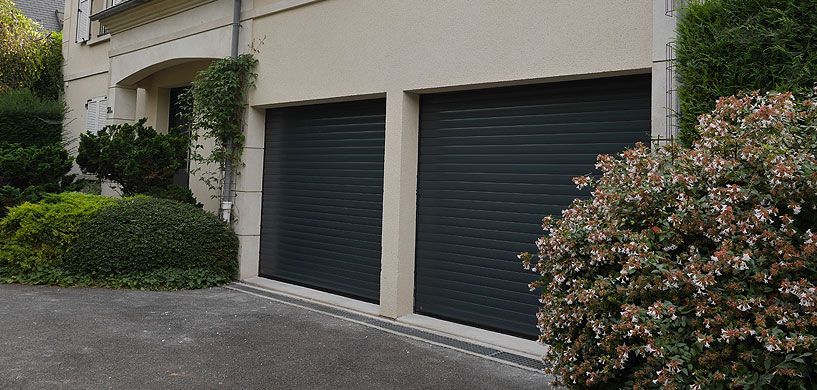 Extérieur de l'appartement avec deux portes de garage foncées, des murs beiges et des arbustes près de l'allée