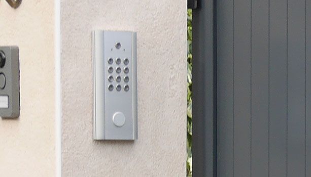 Interphone mural à clavier argenté, à côté d'un portail gris sur un mur extérieur beige