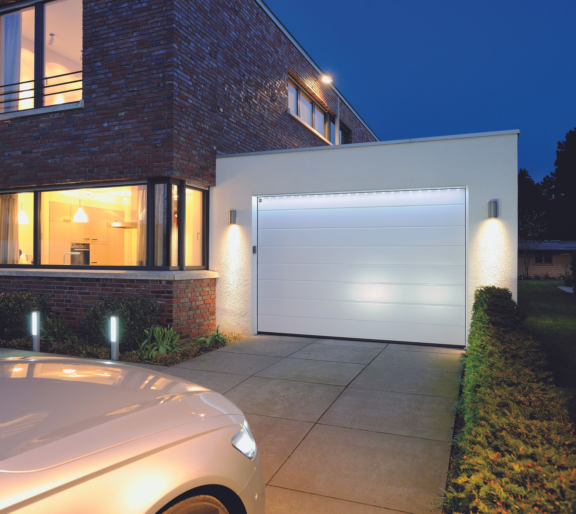 Maison moderne en briques au crépuscule, fenêtres éclairées, porte de garage blanche et allée jouxtant une voiture garée