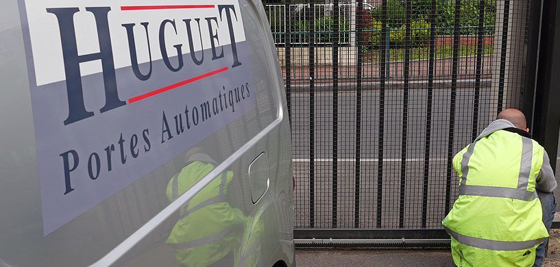 Des ouvriers près d'une clôture, à côté d'un panneau indiquant Huguet Portes Automatiques