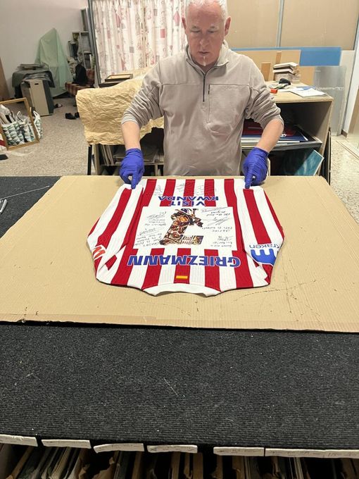Hombre con guantes azules mostrando una prenda a rayas rojas, blancas y azules sobre una mesa en un taller.