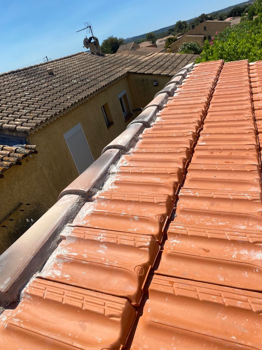 Des tuiles en terre cuite rouge recouvrent un toit en pente, à côté d'une maison beige, sous un ciel bleu azur.