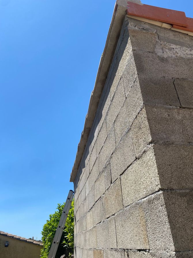 Angle d'un bâtiment en pierre se détachant sur un ciel d'un bleu éclatant, avec des tuiles rouges et une vigne à sa base