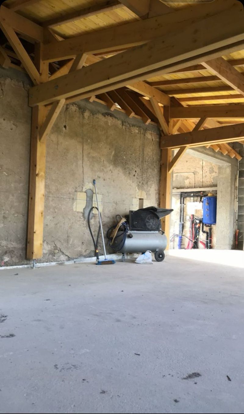 Intérieur d'une vieille grange avec poutres en bois, sol en béton et brouette près d'un mur en plâtre brut.