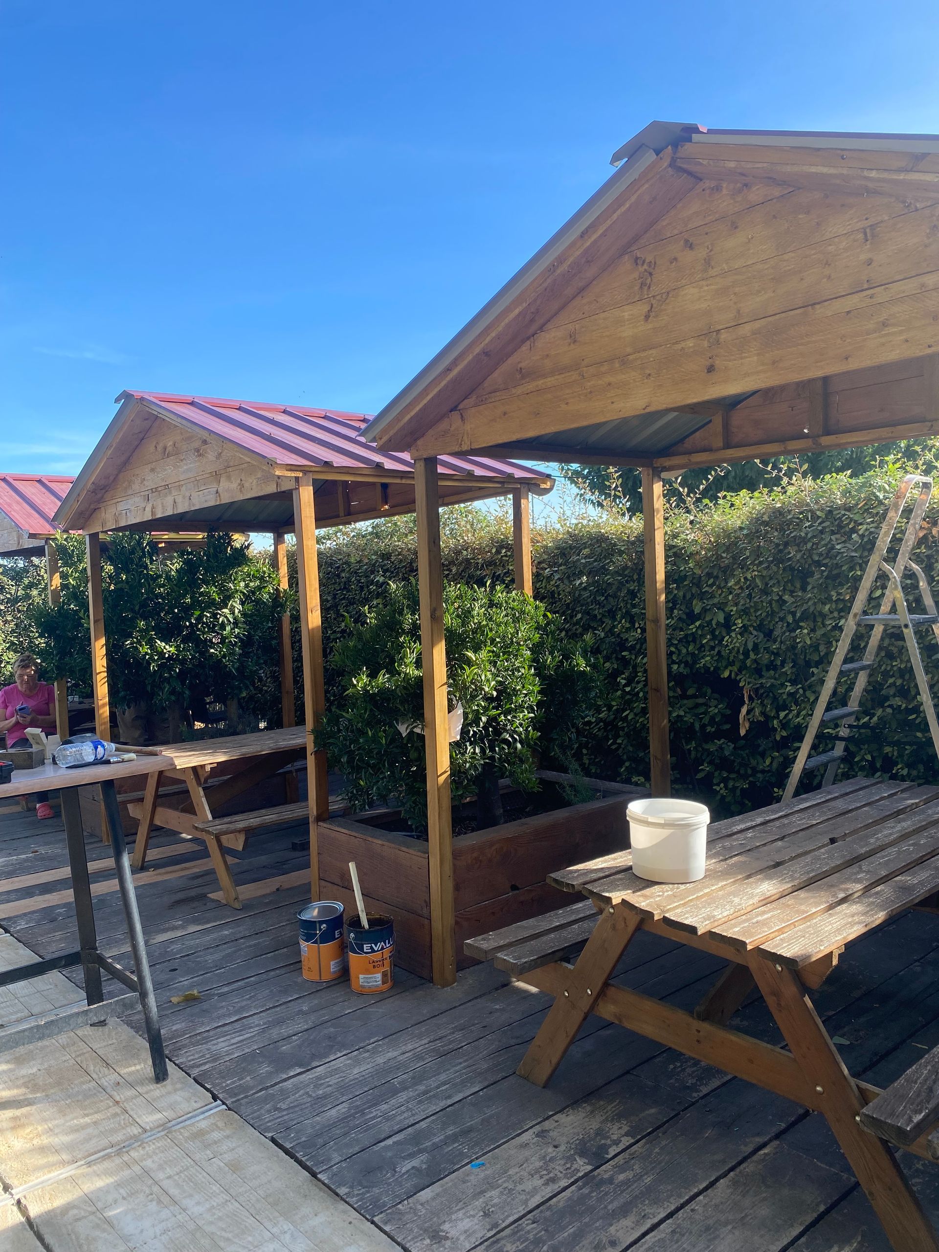 Tables de pique-nique en bois couvertes sous une pergola au toit rouge, sur une terrasse ensoleillée bordée de haies vertes.