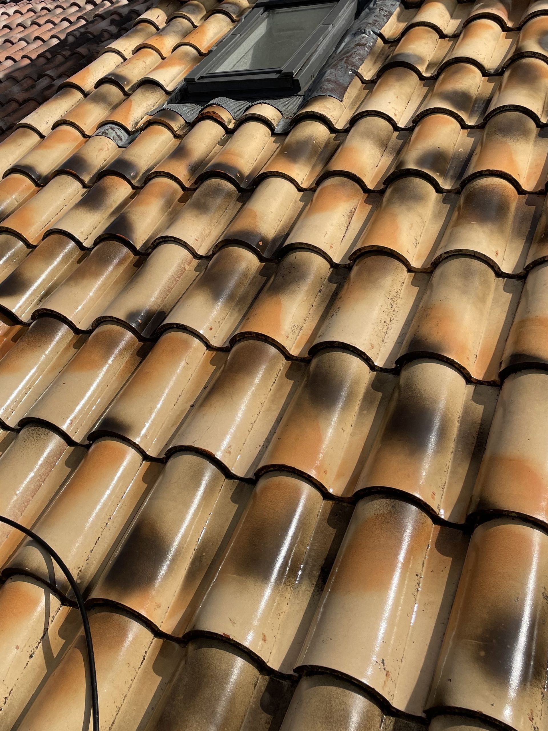 Tuiles de toit en terre cuite avec une petite fenêtre de toit, baignées par la lumière du soleil et marquées de traînées sombres dues aux intempéries