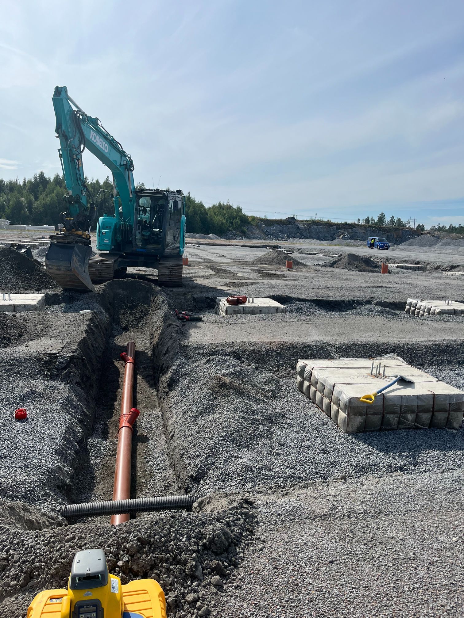 Kaivinkone kaivaa ojaa rakennustyömaalla soralla, putkilla ja betoniperustuksilla.