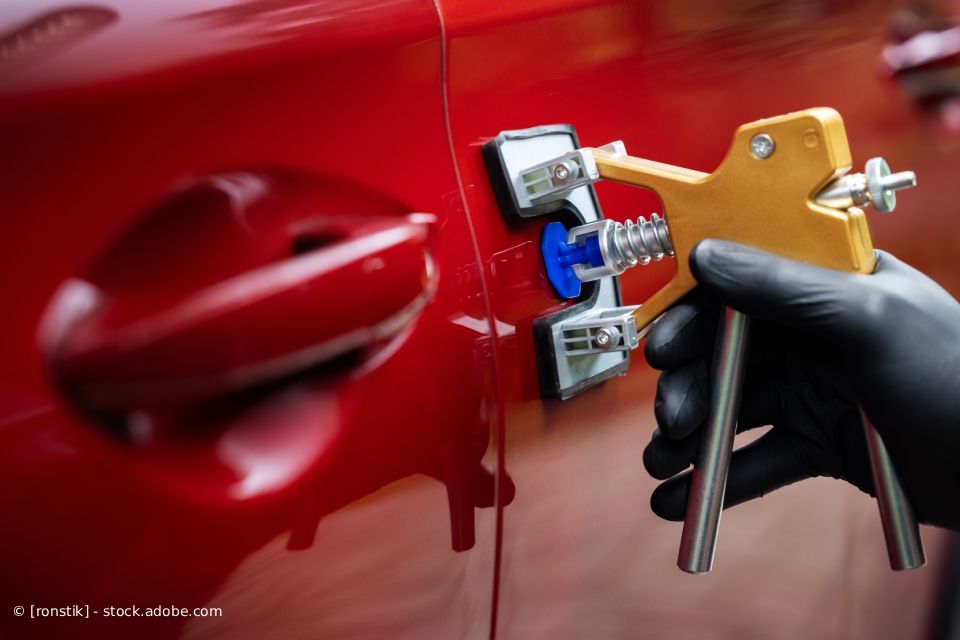 Eine Person mit schwarzen Handschuhen benutzt ein Ausbeulwerkzeug an einer roten Autotür.