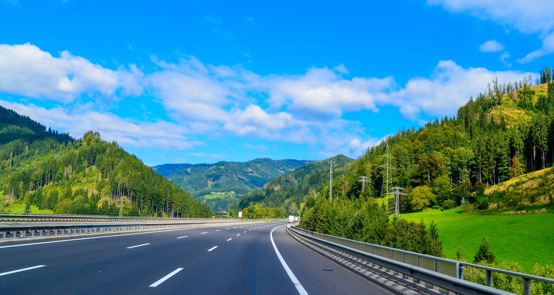 Autoroute déserte serpentant à travers des montagnes verdoyantes et boisées sous un ciel d'un bleu éclatant parsemé de nuages.