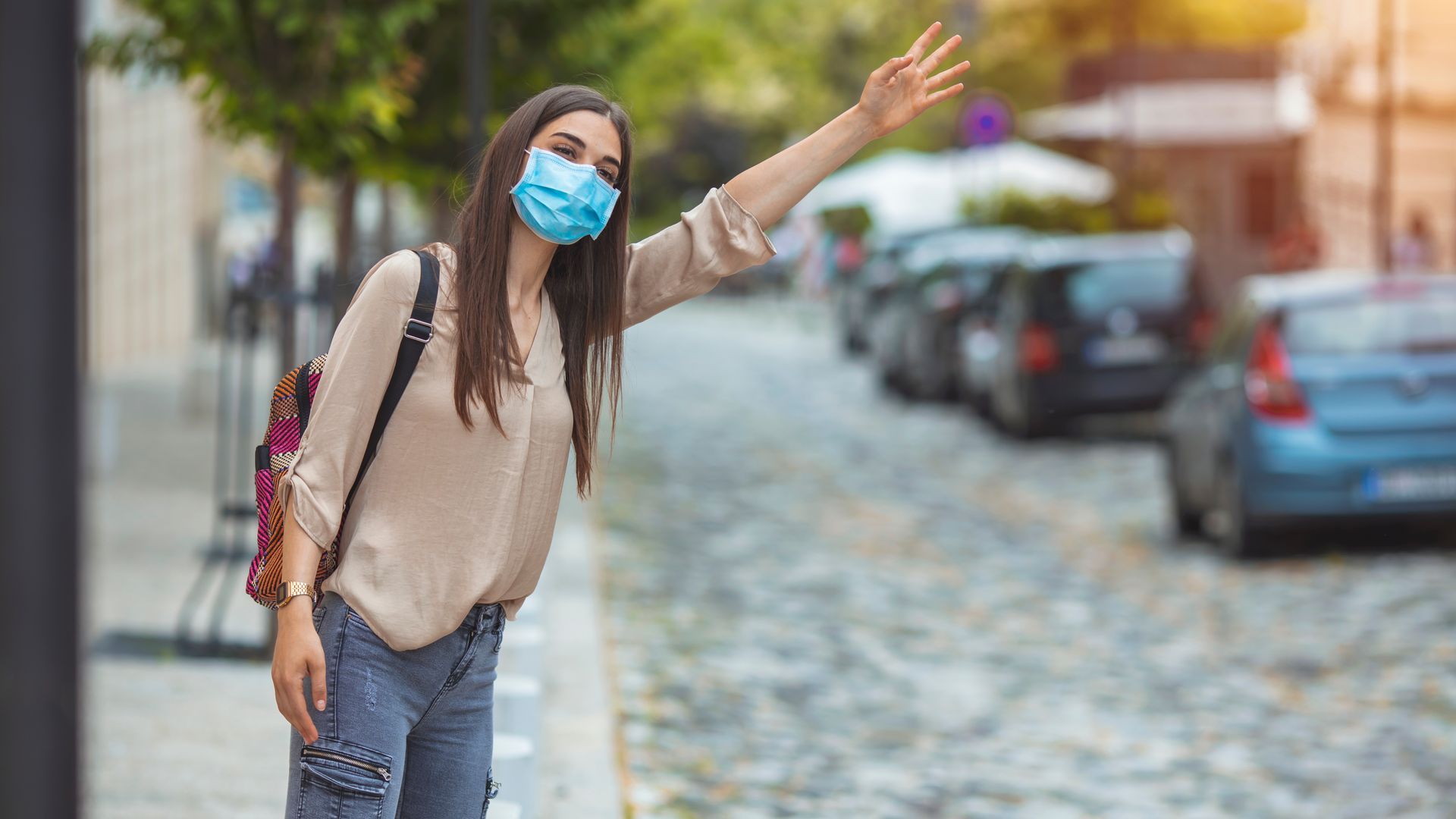 Une personne masquée fait signe de la main dans une rue pavée bordée de voitures garées.