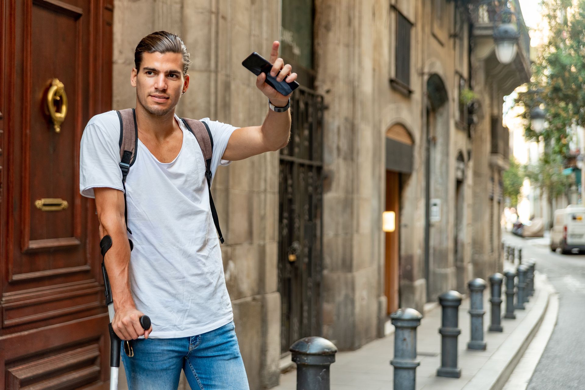 Un homme avec une béquille, se tient debout dans une rue étroite, tenant un téléphone et portant un sac à dos, avec de vieux bâtiments en arrière-plan.