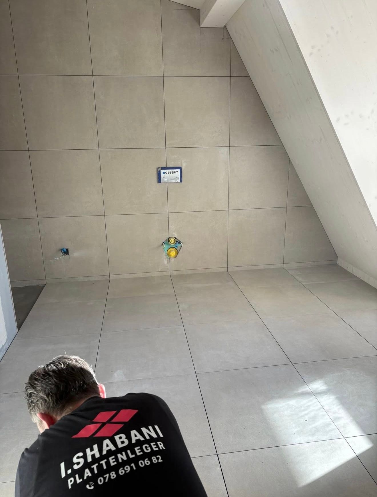 Eine Person mit gelben Handschuhen verlegt mit einem Gummihammer mehrfarbige Pflastersteine auf einem sandigen Untergrund.