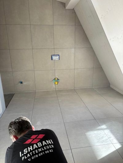 Eine Person mit gelben Handschuhen verlegt mit einem Gummihammer mehrfarbige Pflastersteine auf einem sandigen Untergrund.