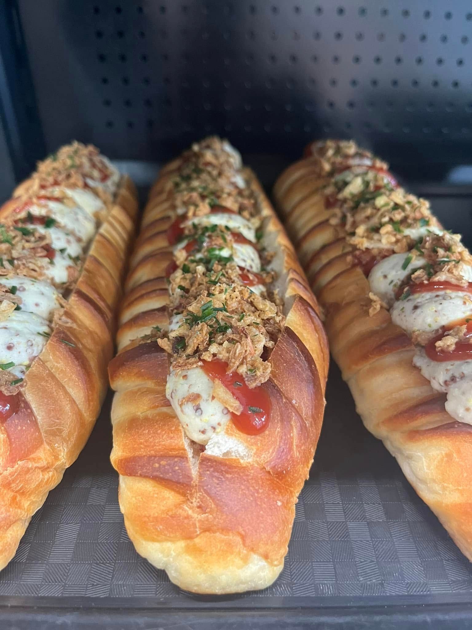 Trois hot-dogs garnis de sauces et d'herbes, alignés sur un plateau sombre.