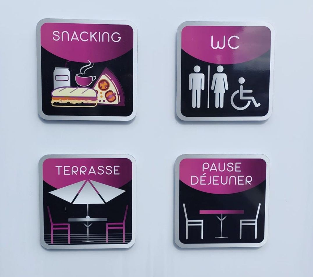 Quatre panneaux muraux roses et noirs indiquent les zones de collation, les toilettes, la terrasse et la pause déjeuner.