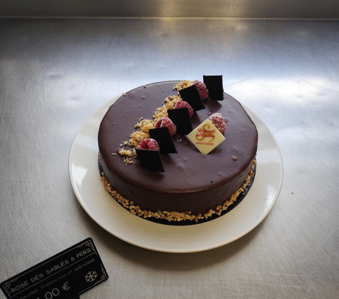 Gâteau au chocolat décoré de formes géométriques en chocolat et de framboises, présenté sur une assiette blanche.