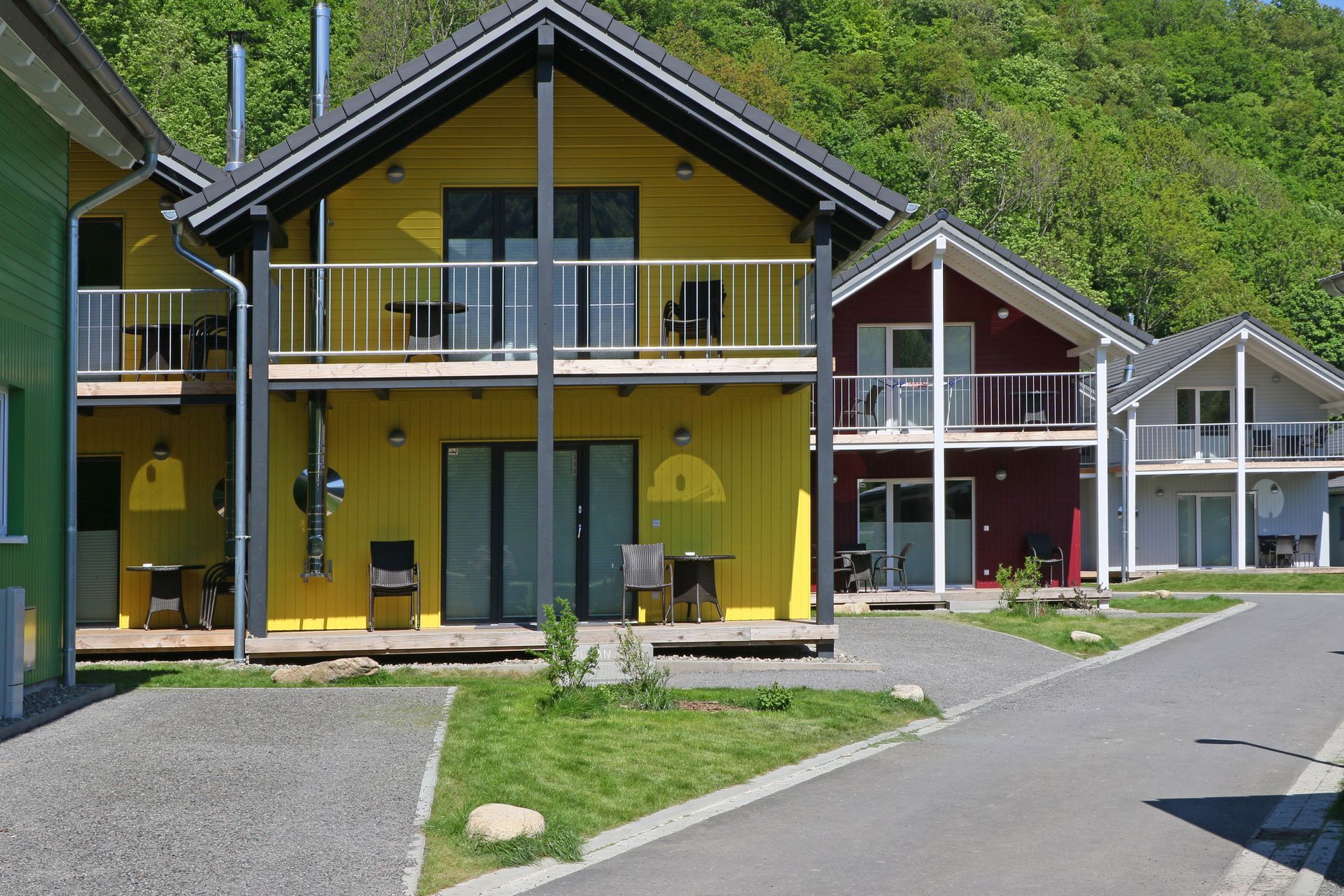 Gelbe und rote Häuser am Seeufer mit Balkonen entlang einer asphaltierten Straße, dahinter grüne Hügel