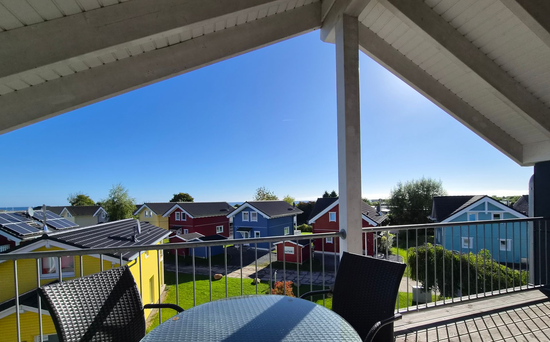 Blick von einer Terrasse auf farbenfrohe Häuser unter einem klaren blauen Himmel, eingerahmt von weißen Dachbalken und einem Geländer.