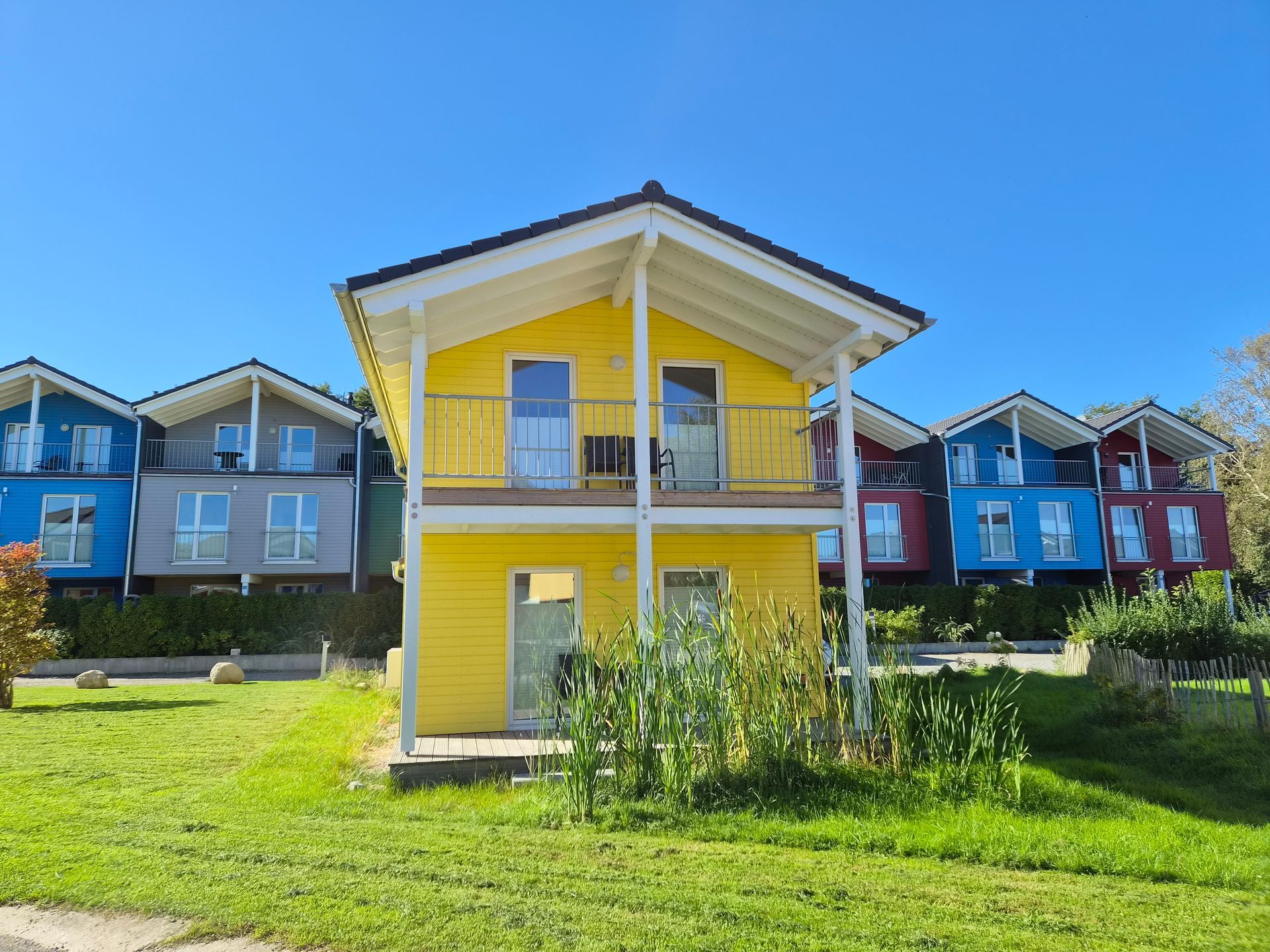Gelbes zweistöckiges Haus mit Balkon, inmitten farbenfroher Häuser auf einem sonnigen grünen Rasen