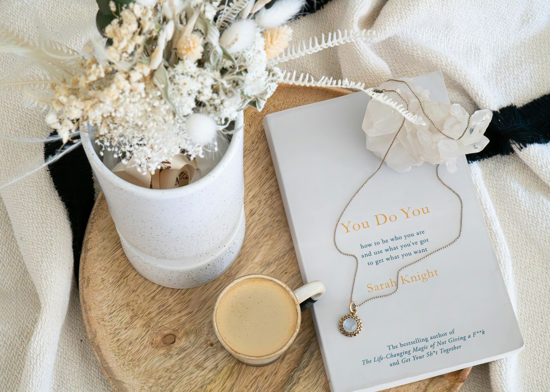 Weißes Trockenblumenarrangement neben einer goldenen Kerze und einer weißen Geschenkbox auf einer gewebten Matte