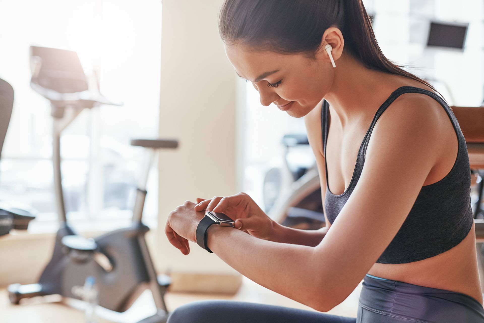 Une personne portant un soutien-gorge de sport et des écouteurs sans fil regarde sa montre connectée, assise dans une salle de sport lumineuse.