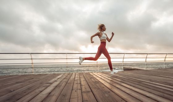 Une coureuse en brassière de sport et legging sprinte sur une promenade en bois au bord de la mer.