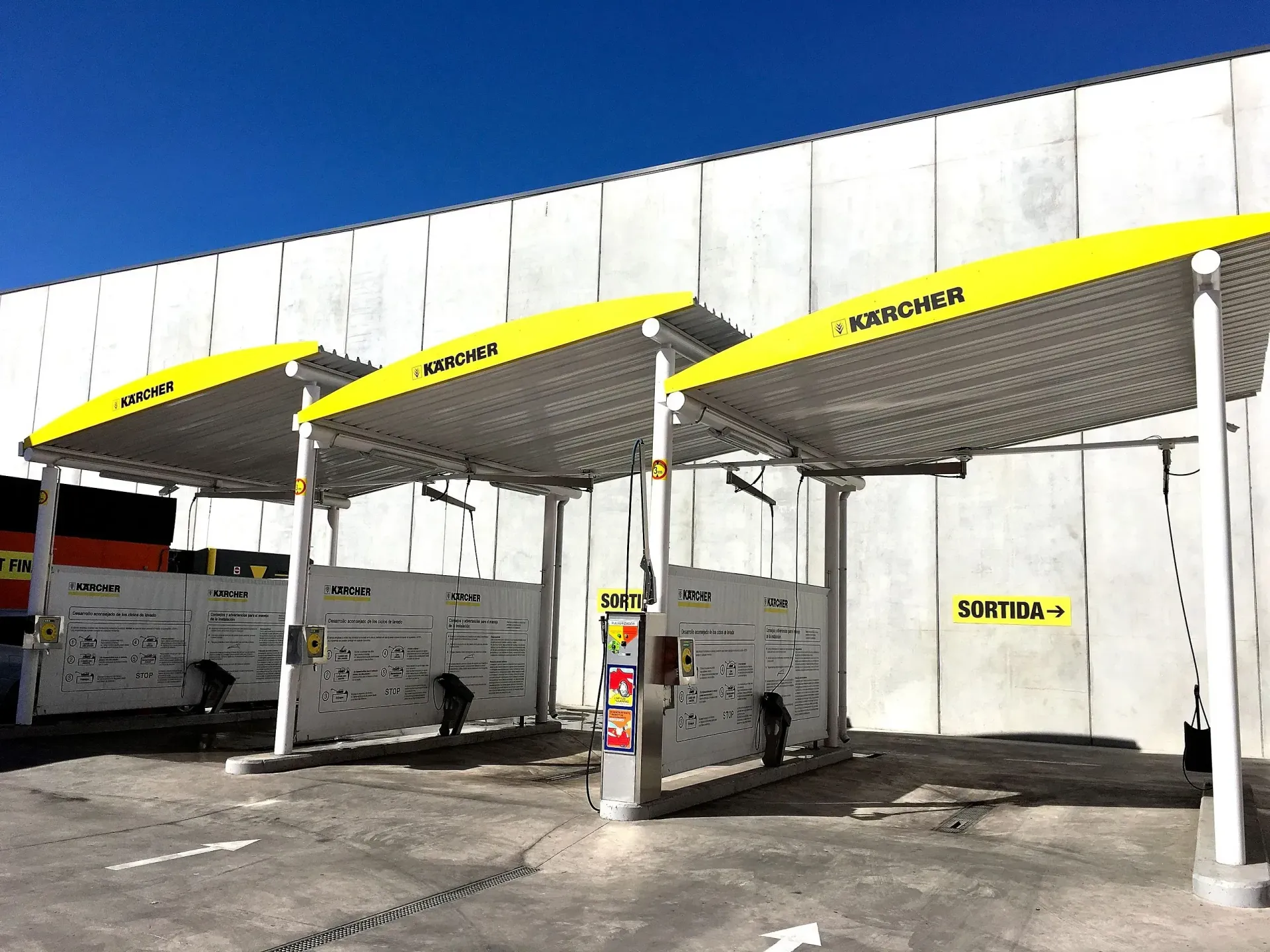 Tres bahías de lavado de vehículos con marquesinas amarillas, paredes de concreto y equipamiento.