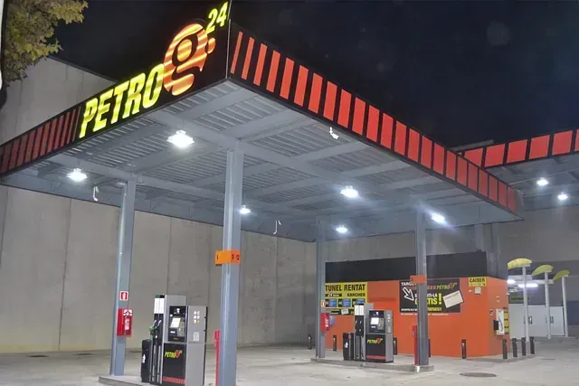 Gasolinera de noche con señalización naranja y negra y surtidores bajo una marquesina.