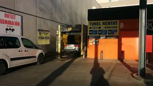 Un coche entra en un túnel de lavado con señalización naranja y amarilla. Hay una furgoneta blanca aparcada cerca.