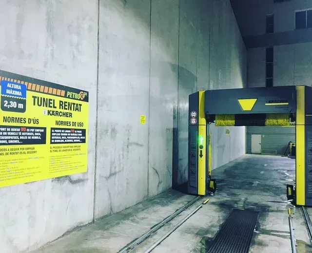 Túnel de lavado de coches, paredes de hormigón gris, maquinaria amarilla y gris, cartel con texto, entorno interior.