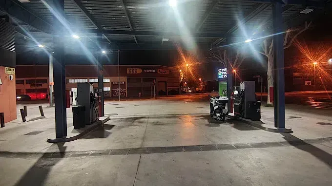 Gasolinera de noche; surtidores iluminados bajo una marquesina. Fachada visible al fondo.
