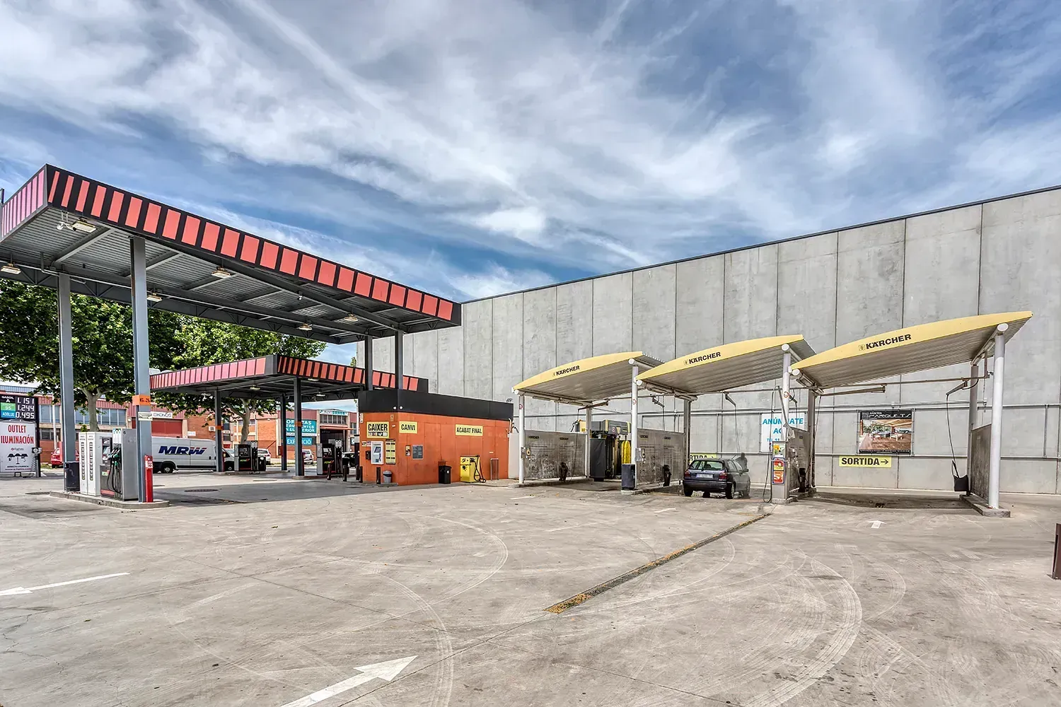 Gasolinera con marquesina roja, zonas de lavado de vehículos y edificio de hormigón.