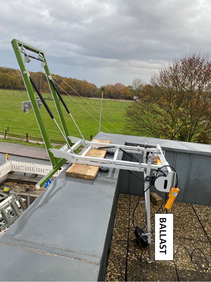 Gerüst auf einem Dach mit einer Winde, Ballast und grünen Stützen vor einem bewölkten Himmel und einer grünen Wiese.