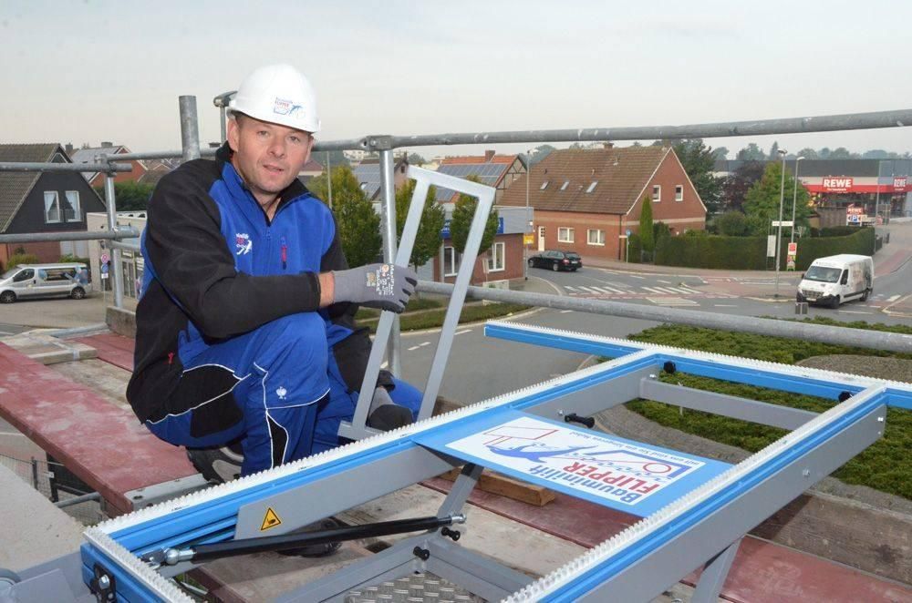 Bauarbeiter kniet auf Gerüst und hält Rahmen fest; blauer Overall, Schutzhelm; Stadtlandschaft im Hintergrund.
