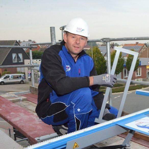 Bauarbeiter auf einem Gerüst, trägt einen weißen Helm und einen blauen Arbeitsanzug und hält einen Metallrahmen.