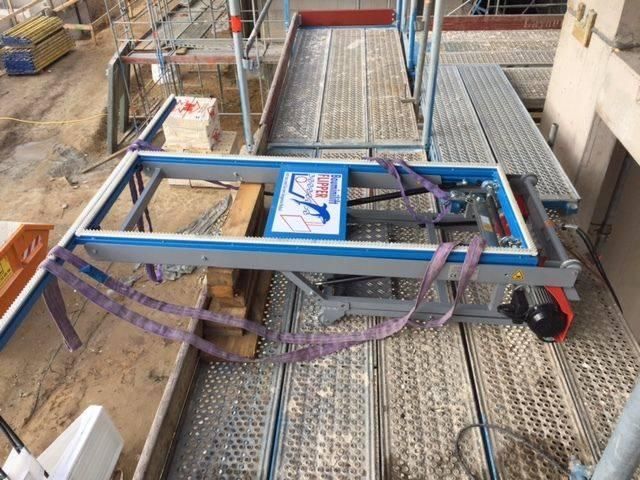 Aufzug auf einer Baustelle; eine Plattform mit Gurten hält einen Fensterrahmen in der Nähe von Baumaterialien.