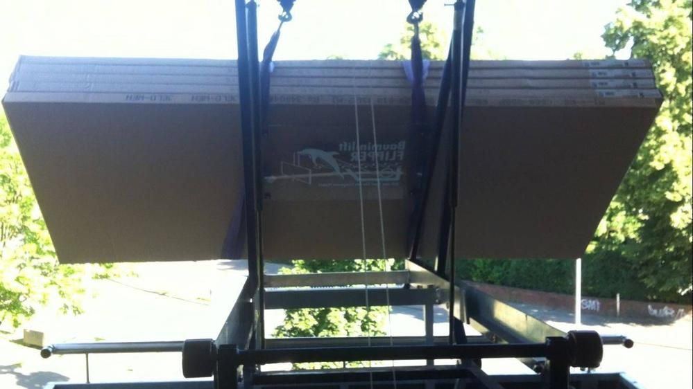 Ein Stapel beigefarbener Trockenbauplatten wird draußen in der Nähe grüner Bäume mit einem Hebezeug angehoben.