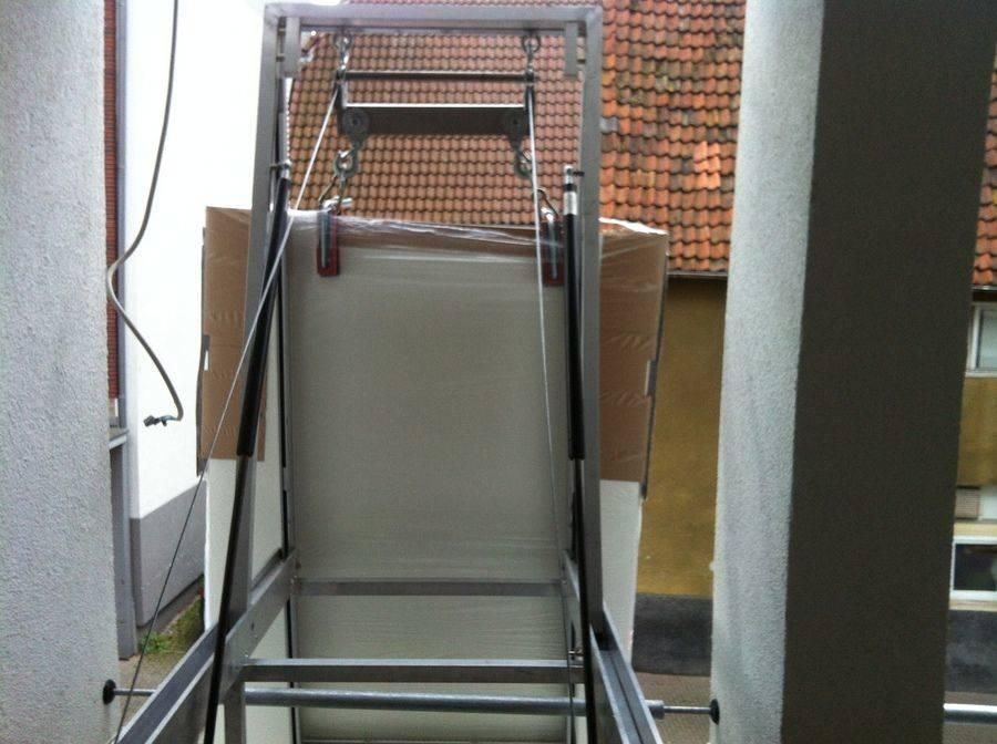 Hebebühne mit einem großen verpackten Gegenstand darauf. Silberner Rahmen, rote Klammern, im Freien.